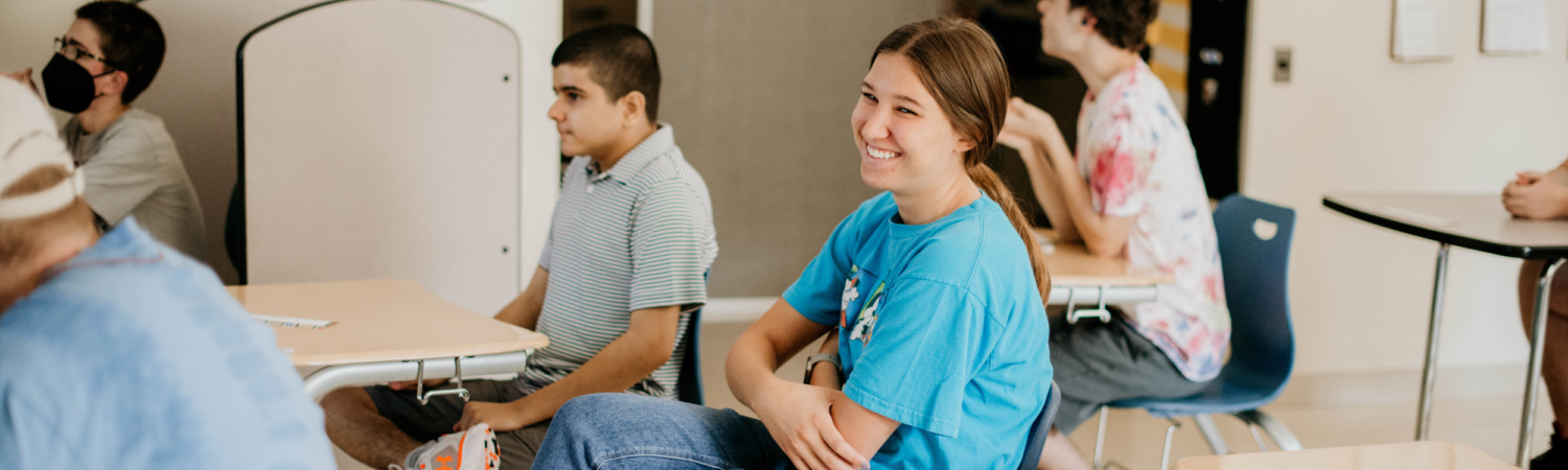 classroom aide sitting at a desk amongst students smiling