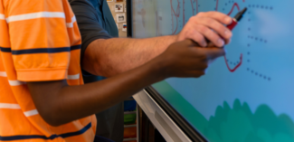 a teacher provides physical prompting to help a student write their name on a SmartBoard - Watson Institute