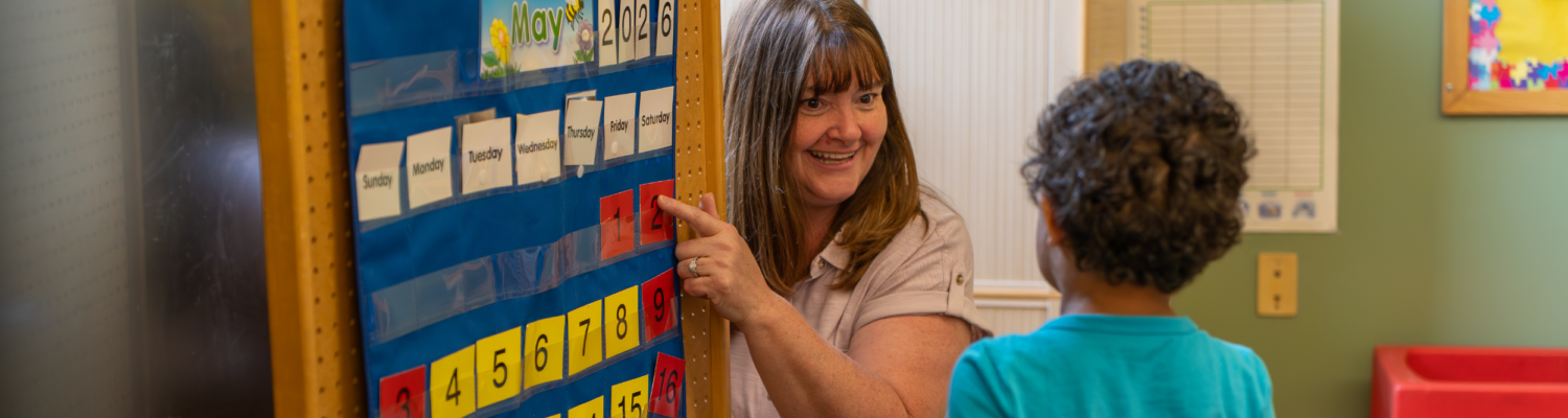 a special education teacher and a preschooler pictured in an inclusive preschool classroom during morning meeting looking at a calendar - Watson Institute