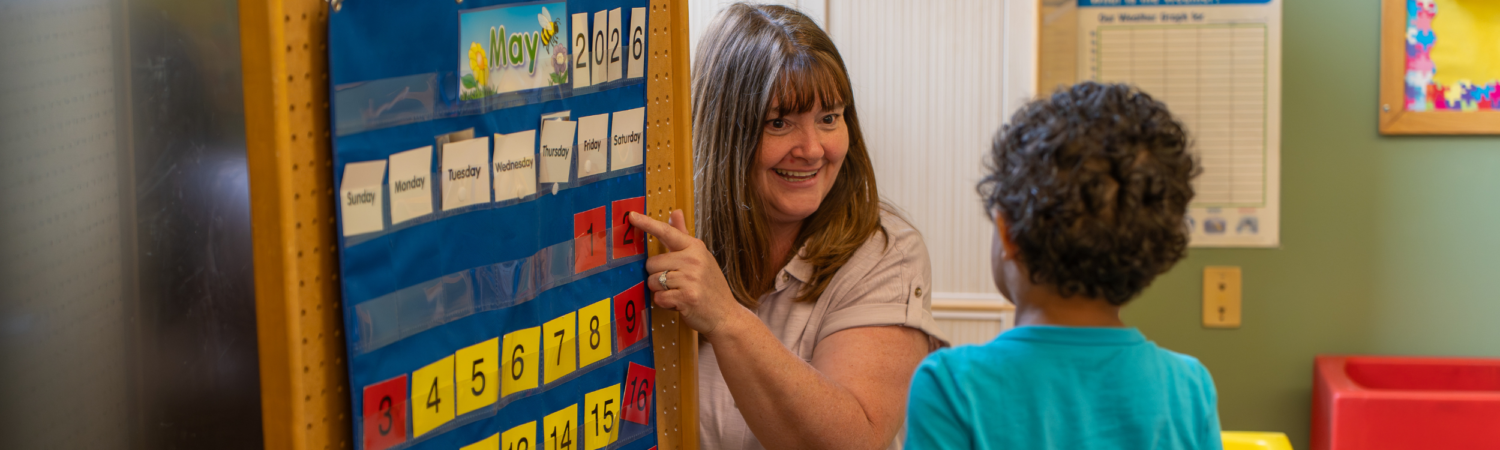 a special education teacher and a preschooler pictured in an inclusive preschool classroom during morning meeting looking at a calendar - Watson Institute