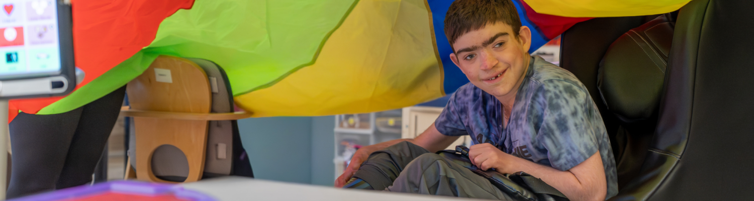 a student with special needs sits under a colorful parachute in his special education classroom - Watson Institute