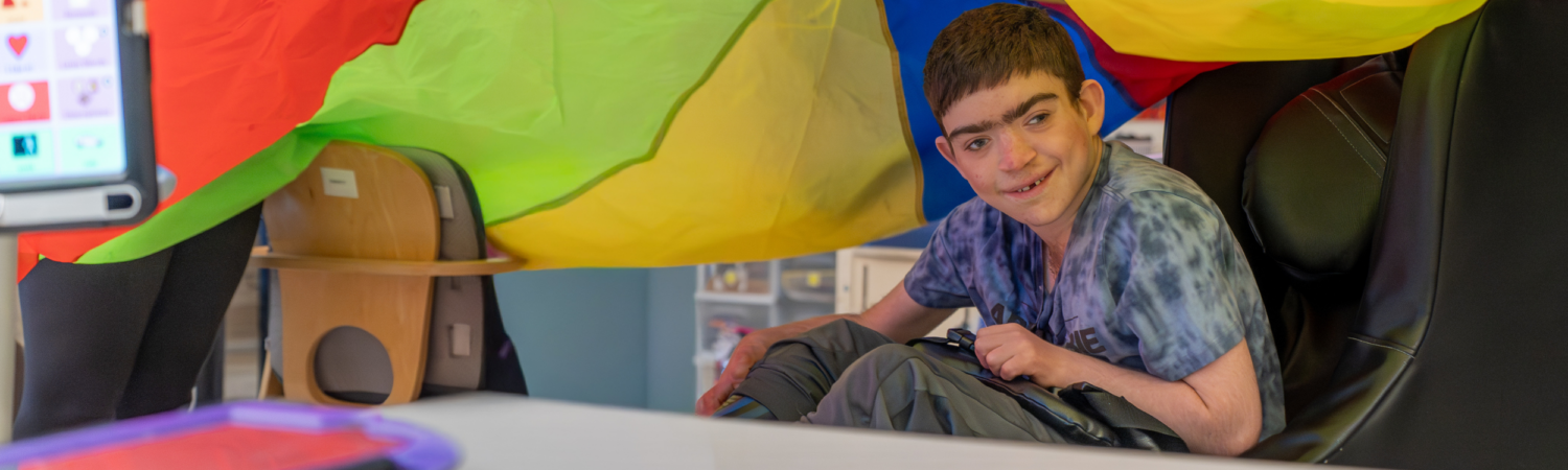 a student with special needs sits under a colorful parachute in his special education classroom - Watson Institute