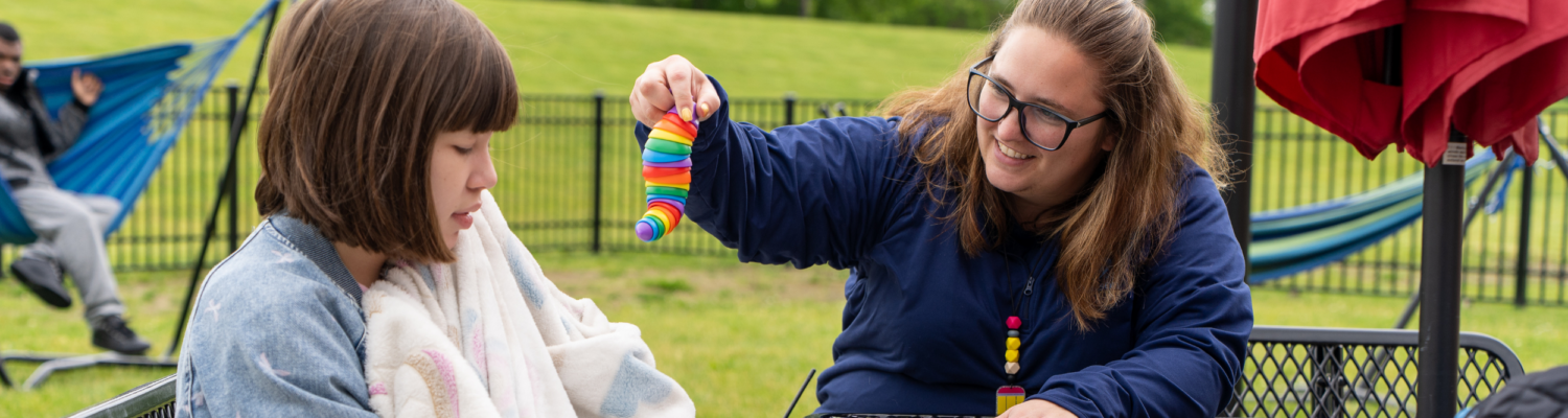special education teacher sits outside with a student with special needs holding a favorite sensory item - Watson Institute