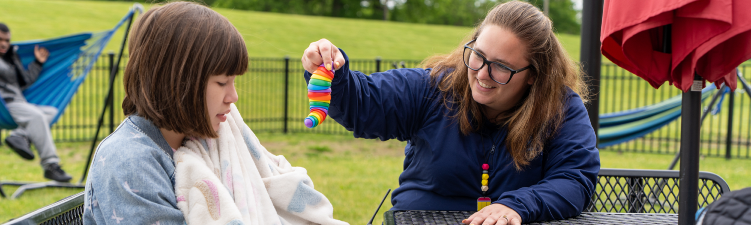 special education teacher sits outside with a student with special needs holding a favorite sensory item - Watson Institute