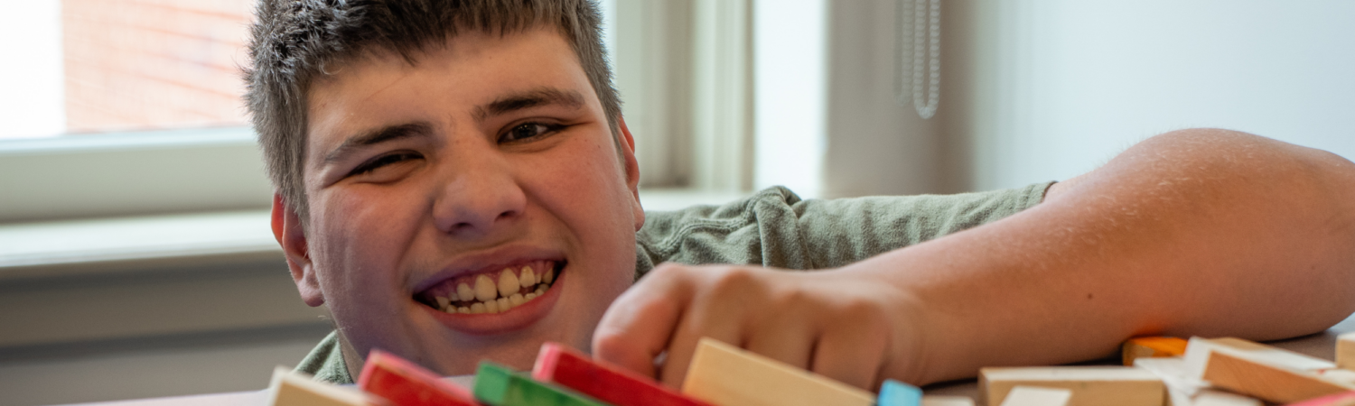 a student with autism is smiling as he plays with dominoes in his classroom - Watson Institute