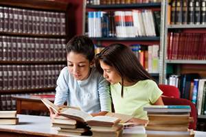 two young girls reading a book in a library at a desk filled with books