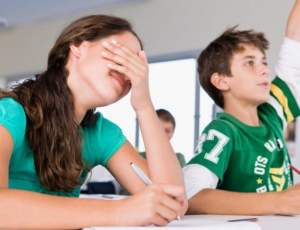 female student seated at desk covering her eyes with her hand while male student next to her is raising his hand