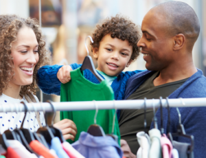A young boy is held by his parents as they look at clothing on a rack of hangers.