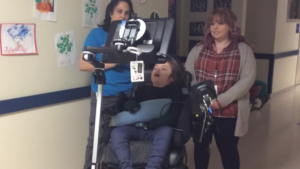 Two educators watch as a student using a power wheelchair drives down the hallway of a school.