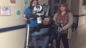 Two educators watch as a student using a power wheelchair drives down the hallway of a school.