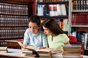two young girls reading a book in a library at a desk filled with books