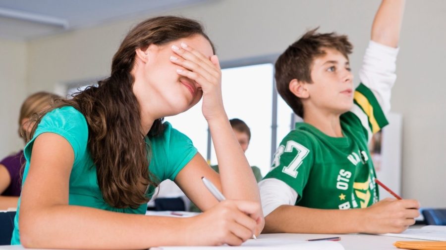 female student seated at desk covering her eyes with her hand while male student next to her is raising his hand