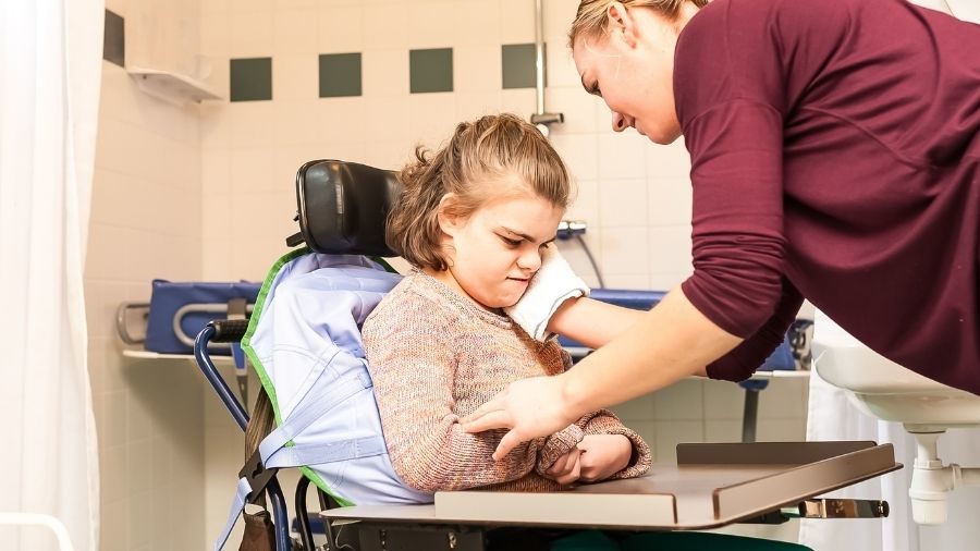 Child with a physical disability seated in a wheelchair while a nurse wipes down her face.