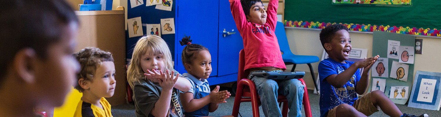 Preschool age students sit on the floor and in chairs in a classroom, clapping along with the teacher.
