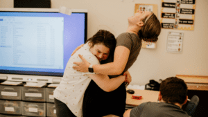 female students hugs educational staff member while she laughs. both are smiling and happy in the classroom