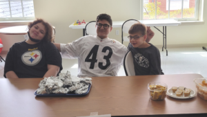 three students with autism seated at a table together smiling at the camera while selling hot pretzels