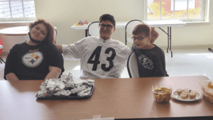 three students with autism seated at a table together smiling at the camera while selling hot pretzels