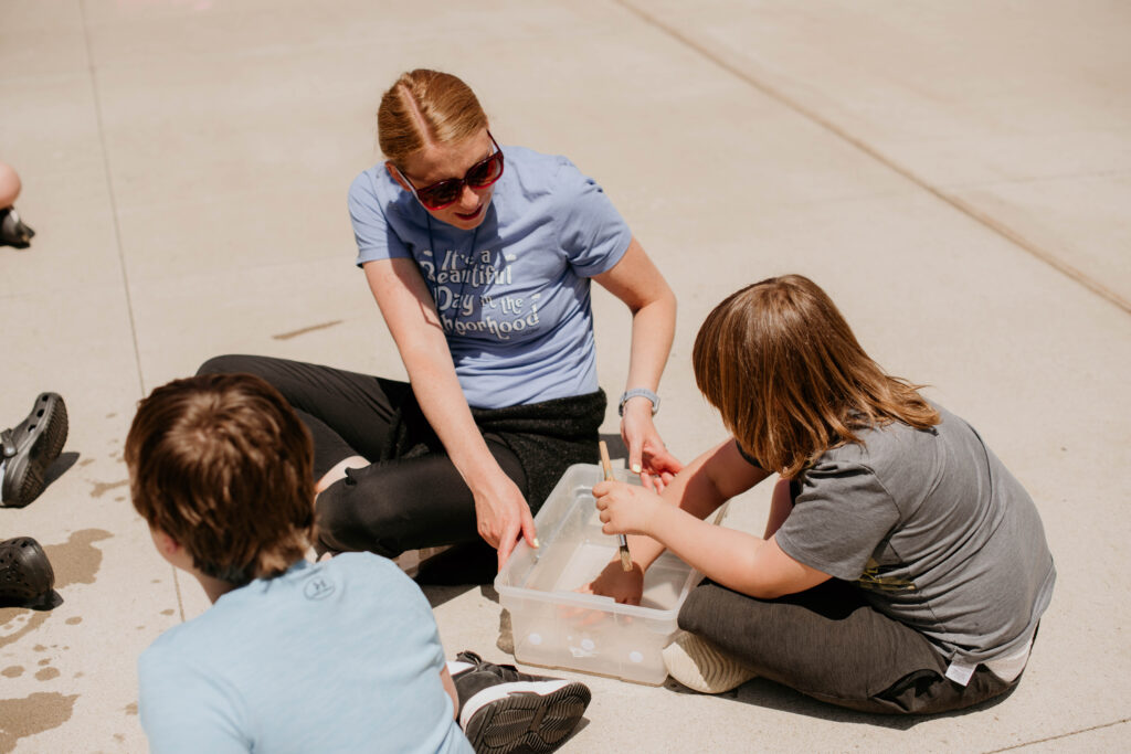Teacher is sitting on the ground outside with students during the summer school program doing sensory activities.
