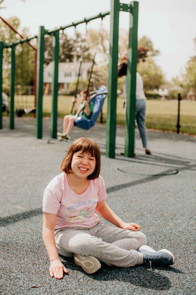 A student is pictured sitting on the ground of the playground smiling. In the background a student is being pushed in an adapted swing by a teacher.