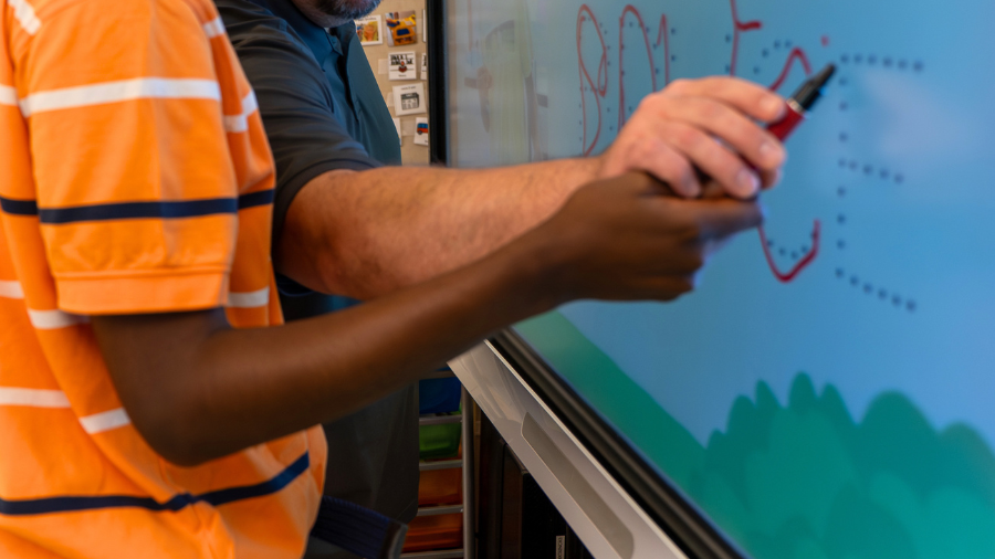 a teacher provides physical prompting to help a student write their name on a SmartBoard - Watson Institute